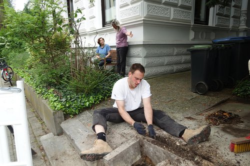 In the background, two women are talking and pointing at a person doing gardening work in the foreground.