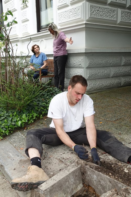 In the background, two women are talking and pointing at a person doing gardening work in the foreground.