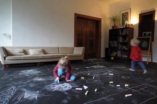 Two children colour a floor covered with black paper in a living room.