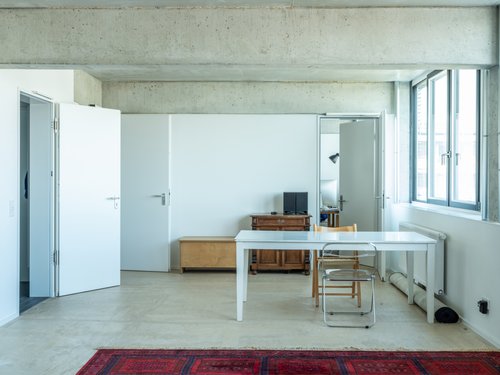 White room, known as the Nukleuswohnen, with numerous doors and a glazed façade as well as simple furnishings.